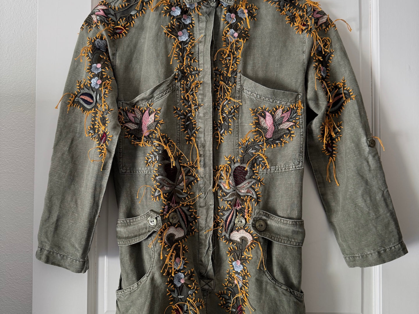 An olive green denim jacket with intricate multicolor floral embroidery, hanging on a wooden hanger against a white paneled door.