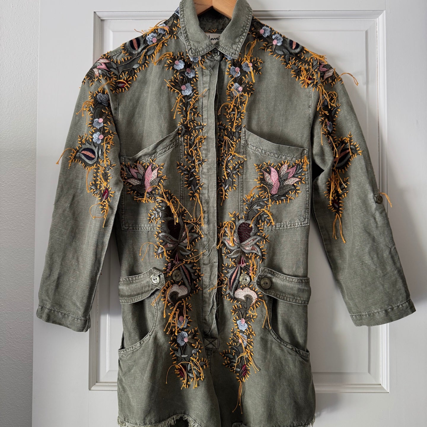 An olive green denim jacket with intricate multicolor floral embroidery, hanging on a wooden hanger against a white paneled door.
