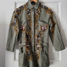 An olive green denim jacket with intricate multicolor floral embroidery, hanging on a wooden hanger against a white paneled door.