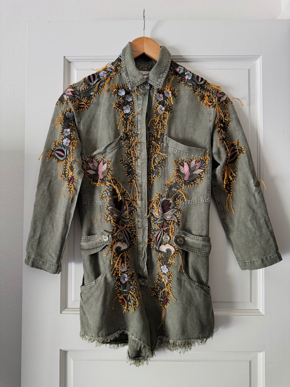 An olive green denim jacket with intricate multicolor floral embroidery, hanging on a wooden hanger against a white paneled door.