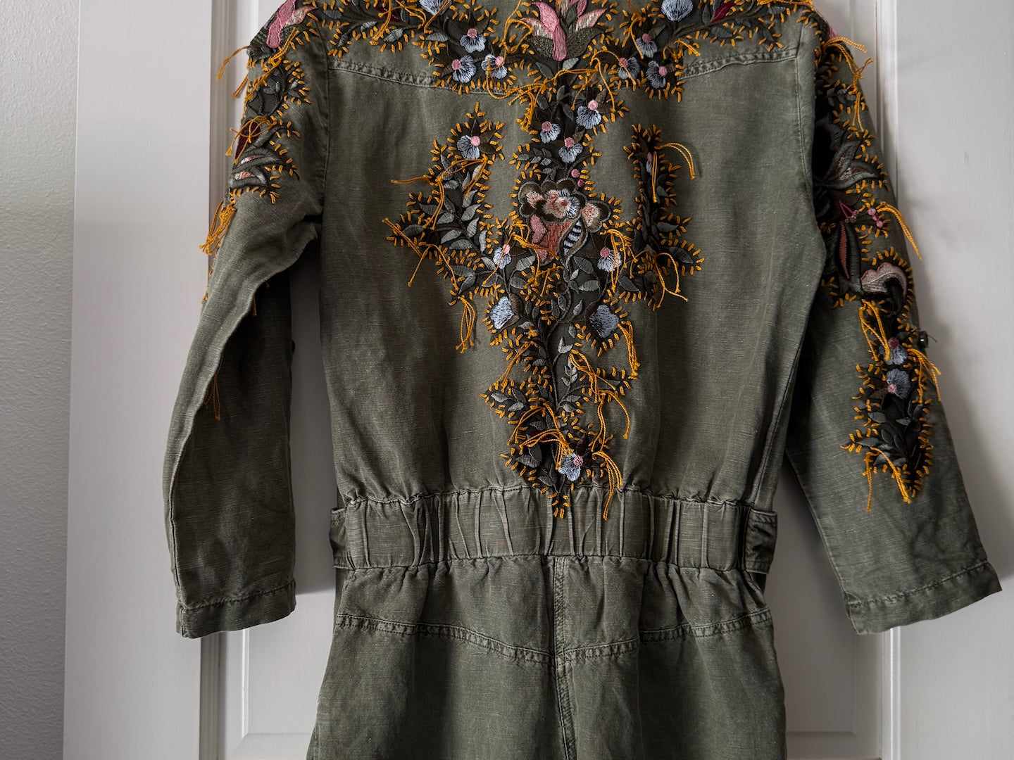 An olive green denim romper with extensive colorful floral embroidery on the back, hanging on a wooden hanger against a white paneled door.