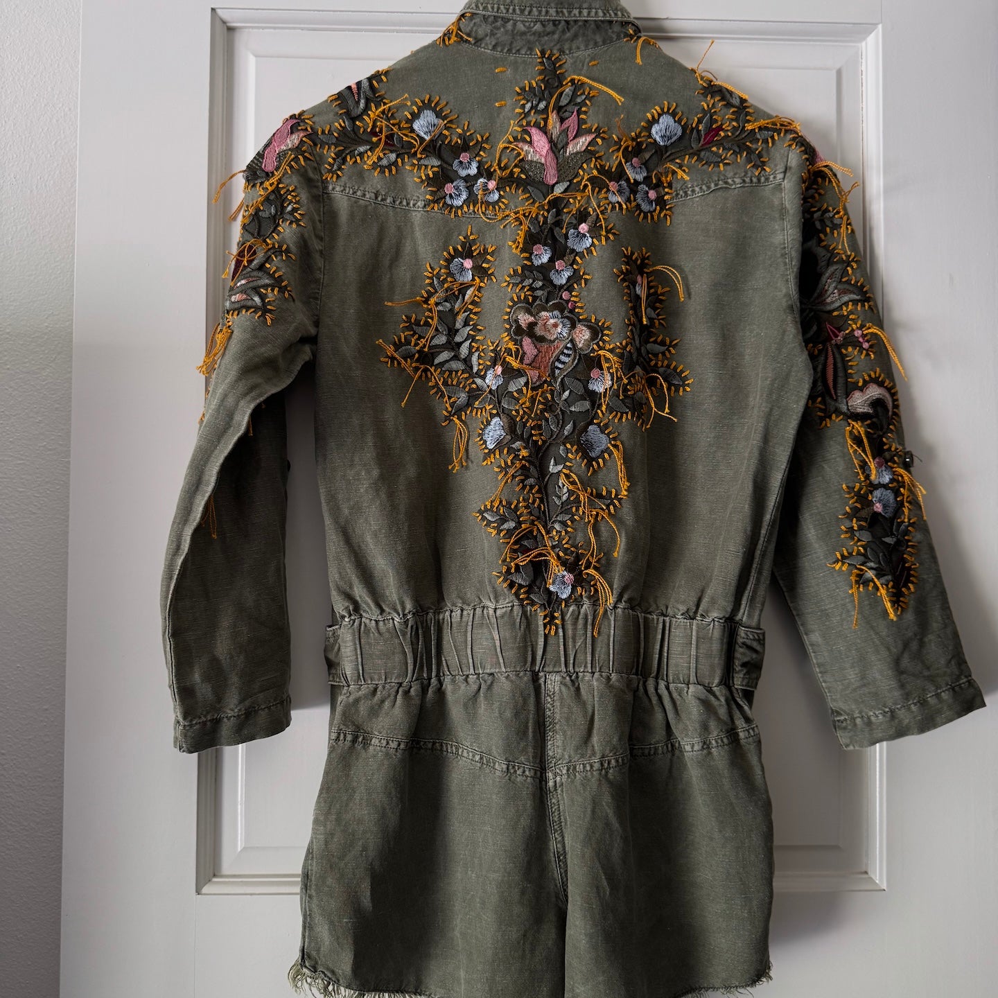 An olive green denim romper with extensive colorful floral embroidery on the back, hanging on a wooden hanger against a white paneled door.