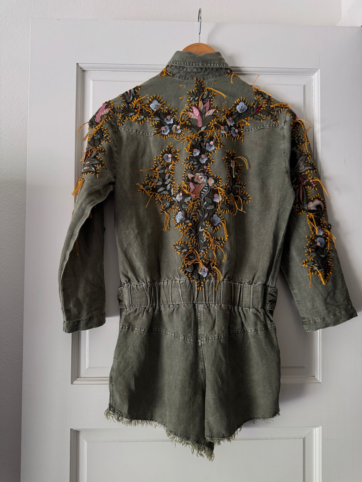 An olive green denim romper with extensive colorful floral embroidery on the back, hanging on a wooden hanger against a white paneled door.